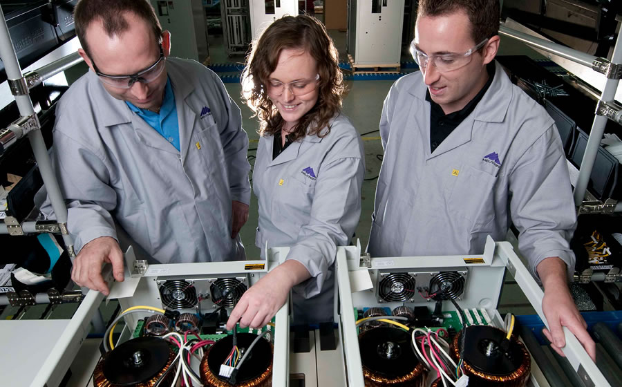 Melitron employees with safety glasses inspect the electro-mechanical assembly within a metal enclosure.