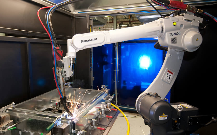 A robotic welding arm makes sparks fly in a welding cell as part of the fabrication of a metal enclosure.